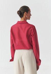 Chemise en coton rouge, col, avec des manches longues. Présente une coupe décontractée et des poignets à boutons cachés, portée avec un pantalon de couleur claire.