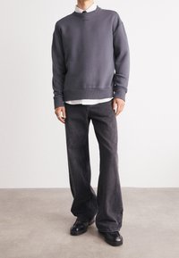 Un sweat-shirt gris à col rond avec des poignets côtelés, superposé sur une chemise blanche à col. Assorti avec un jean noir à jambes larges et des chaussures noires.