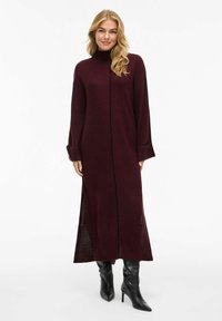Abito lungo color marsala con texture a coste, colletto alto e spacchi laterali. Presenta un taglio dritto e maniche lunghe, abbinato a stivali neri con tacco alto.