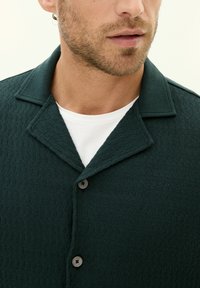 Tummanvihreä tekstuurinen paita, jossa on leveä kaulus, kolme nappia ja valkoinen sisäpaita. Kankaan pinnassa on hienovarainen kuvio.