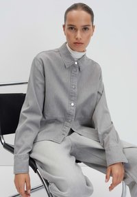 Gray denim jacket with a button-up front, pointed collar, and long sleeves. Paired with matching gray pants, sitting on a black chair.
