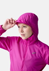 Fuchsia lightweight jacket with a fitted hood, zip closure, and elastic cuffs. Reflective logo accent on the upper left chest.