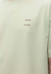 Ανοιχτό μπέζ T-shirt με δύο λογότυπα "SAMSØE" τυπωμένα με μικρή απλή γραμματοσειρά στο πάνω μέρος του στήθους.