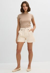 Camiseta ajustada de color beige claro combinada con pantalones cortos de lino crema que cuentan con dos bolsillos frontales y cierre de doble botón. Zapatos planos de tejido beige completan el look.