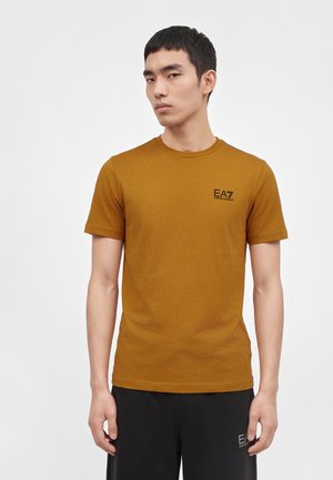 Homme portant un t-shirt à manches courtes marron moutarde avec un petit logo noir EA7 Emporio Armani sur la poitrine et un pantalon noir.