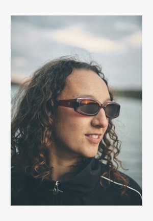 Sötét lencsés napszemüveg bordó kerettel, elegáns kialakítással és ívelt szárakkal. Egy göndör hajú személy viseli.