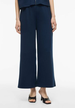Personne portant un pantalon large bleu marine et des mules à talons ouvertes noires, debout devant un fond blanc.