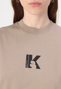 Bež majica iz bombaža z črnim grafičnim logotipom, ki vsebuje silhueto in črko "K". Okrogli ovratnik in kratki rokavi.
