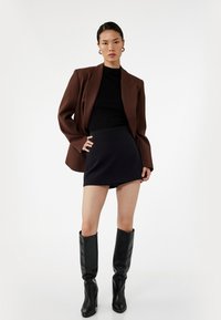 Bruine oversized blazer, zwarte fitted coltrui, marineblauwe minirok en zwarte kniehoog laarzen. Gladde texturen en contrasterende kleuren.