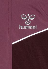 Tessuto atletico color burgundy con striscia accentata bianca. Presenta un logo Hummel ricamato con un design simmetrico. Texture liscia.
