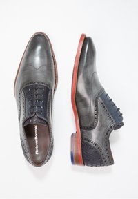 Scarpe in pelle grigia con un design brogue e accenti in tessuto blu navy testurizzato. Suola rossa ben visibile e cuciture dettagliate lungo i bordi.