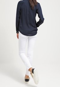 Mörkblå långärmad blus, vita slim-fit jeans och avslappnade svarta och beige sneakers med en strukturerad sula. Blusen har en rundad nederkant.
