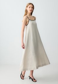 Maxi dress di lino beige chiaro con sottili spalline nere, vestibilità rilassata, silhouette fluida e spacchi laterali, indossato con sandali neri con tacco.