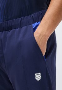 Χέρι που εισέρχεται στην τσέπη σκουρόμπλε παντελονιού με ελαστική μέση, εμφανίζοντας μπλε εσωτερική επένδυση και το λογότυπο K-Swiss στον μηρό.