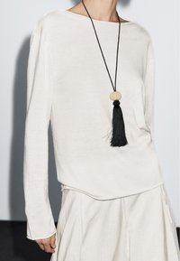 Licht beige longsleeve top van zachte stof, met een ronde halslijn. Gecombineerd met een vloeiende, lichtgekleurde rok en een zwarte ketting met kwastje.