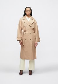 Μπεζ παλτό-καπαρντίνα με διπλό πέτο, μεγάλους γιακάδες και ζώνη στη μέση που δένει. Ομαλή υφή, με μακριά μανίκια και πλαϊνές τσέπες.