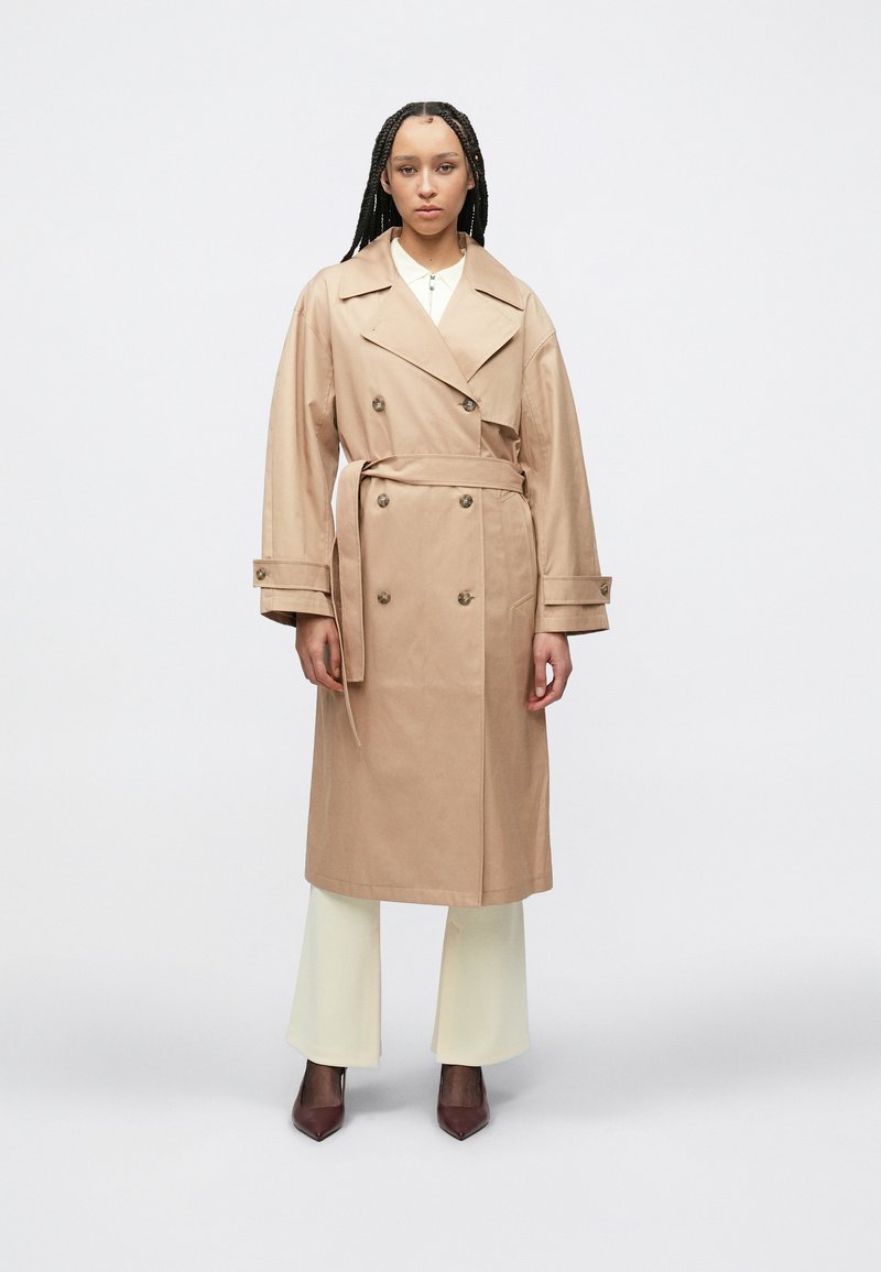 Μπεζ παλτό-καπαρντίνα με διπλό πέτο, μεγάλους γιακάδες και ζώνη στη μέση που δένει. Ομαλή υφή, με μακριά μανίκια και πλαϊνές τσέπες.