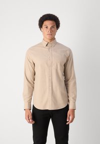Camicia beige a maniche lunghe con colletto button-down, taschino sul petto e trama sottile, abbinata a pantaloni neri.