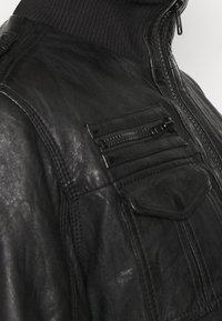 Zwarte leren jas met een gestructureerde afwerking, voorzien van een hoge kraag, ritssluiting en meerdere zakken met ritsen en kleppen.