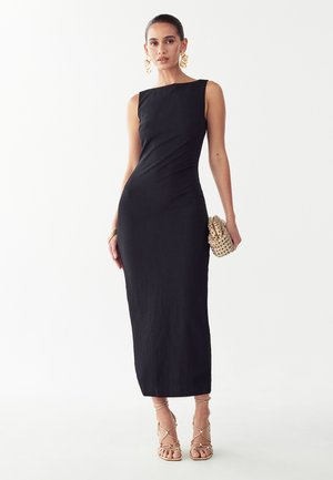 Vrouw draagt een zwart mouwloos midi-jurk met gouden sandalen met bandjes, grote gouden oorbellen, armbanden en houdt een geweven gouden clutch vast.