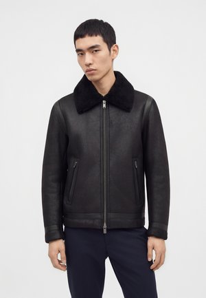 Jeune homme portant une veste en cuir noire avec une fermeture éclair devant et un col en fausse fourrure noire, debout devant un fond clair uni.