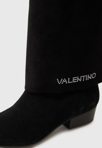 Bottes noires en daim à mi-mollet avec un talon bloc, dotées d'un revers et ornées de strass formant le mot "VALENTINO."