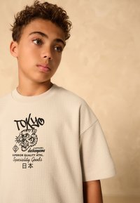 Maglietta a maniche corte color beige chiaro con motivo a griglia testurizzato. Presenta una grafica nera di una tigre e testo giapponese sul davanti.