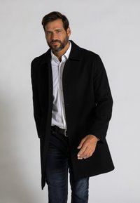 Abrigo de lana negro con cuello, cierre de botones y bolsillos laterales, llevado sobre una camisa blanca y jeans azules. Textura suave, corte entallado.