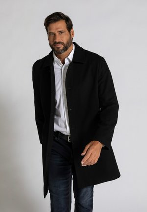 Cappotto di lana nero con collare, chiusura a bottoni e tasche laterali, indossato sopra una camicia bianca e jeans blu. Tessuto liscio, vestibilità sartoriale.