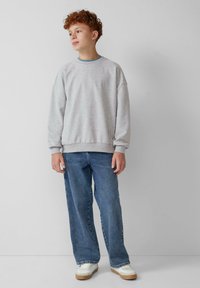 Felpa grigia con una vestibilità rilassata, abbinata a jeans blu e sneakers bianche con suole in gomma. Design semplice, texture morbida.