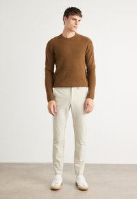 Sweater castanho de malha com gola redonda, calças beiges de corte ajustado e ténis brancos com solas castanhas claras. Textura suave, design ajustado.