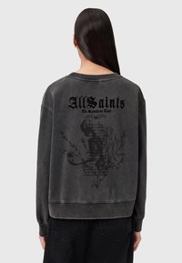 Una persona con lunghi capelli scuri indossa un felpa nera sbiadita di AllSaints che presenta un design con scheletro e fiori sulla schiena.