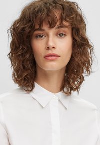 Bílá košile s límečkem a knoflíkovým zapínáním vyrobená z hladké látky. Modelka má kudrnaté vlasy a přirozený makeup, který zdůrazňuje přirozený vzhled.