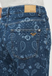 Μπλε denim τζιν με ανοιχτά λουλουδένια και παίζει σχέδια, που διαθέτουν πίσω τσέπη με καφέ δερμάτινη ετικέτα λογότυπου Roxy και θηλιά ζώνης.