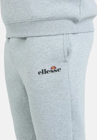 Grijze sweatpants gemaakt van zachte, textuurstof met een zijzak en een zwart "ellesse" logo met daarboven een kleurrijke zonafbeelding.