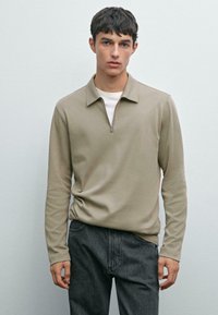 En ung man som bär en beige zip-up långärmad tröja över en vit undertröja och svarta jeans, står mot en ljusgrå vägg.