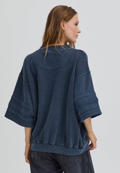 Femme aux longs cheveux portant un sweat-shirt à manches courtes bleu foncé surdimensionné et un jean noir, vue de dos sur un fond uni.