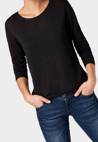 Person in einem strukturierten schwarzen Langarmoberteil und blauen Jeans, mit den Händen in den Taschen, stehend vor einem einfarbigen Hintergrund.