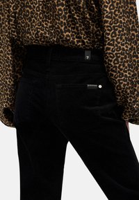 Pantalones vaqueros de pana negros con una cintura alta y bolsillos traseros. Acentuados con una etiqueta de la marca en la cintura y un modelo que lleva una blusa de estampado de leopardo.