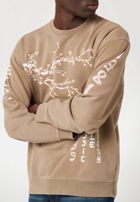 Rudas sportinis džemperis su rožinės spalvos gėlių raštu priekyje ir baltu tekstu ant rankovių. Apvalus kaklas ir guminiai rankogaliai.