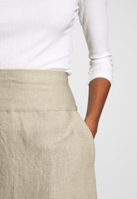 Beige linnekjol med bred midjeband, som har två sidfickor och en lätt skrynklig textur. Ribbad vit topp delvis synlig.