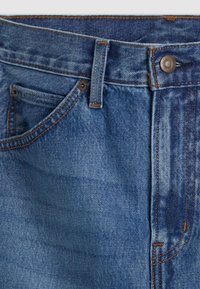 Gros plan sur un jean en denim bleu montrant la poche avant, la boucle de ceinture, le bouton et les détails de couture orange.