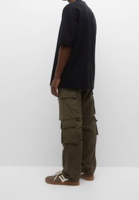 T-shirt noir oversize, pantalon cargo vert avec multiples poches, et baskets marron avec des accents blancs. Tissu lisse avec une coupe décontractée.