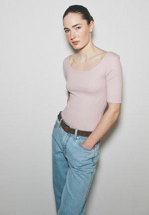 Žena s tmavými vlasmi zviazanými dozadu, oblečená v svetloružovom tričku s výstrihom do U a voľných svetlomodrých džínsoch s hnedým opaskom, stojaca s rukami vreckách.