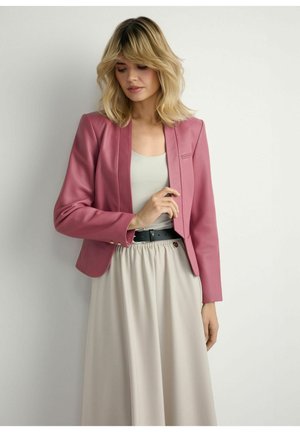 Blondynka ubrana w różowy żakiet, białą bluzkę, beżową spódnicę i czarny pasek, stojąca na tle gładkiej białej ściany.