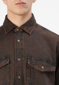 Close-up of a person wearing a faded black denim jacket with brown stitching, buttoned front, and two chest pockets with buttons.
