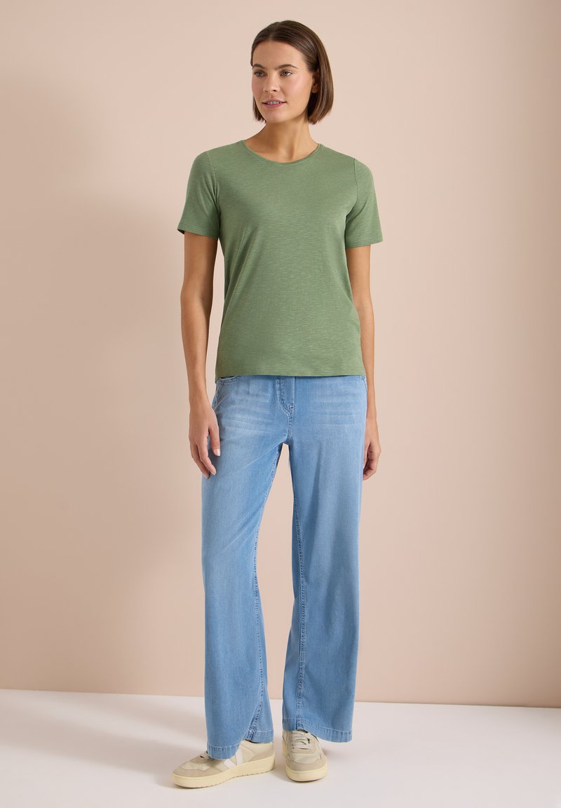 Femme debout portant un t-shirt vert clair à manches courtes, un jean large bleu clair et des baskets beiges sur un fond beige uni.