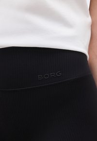 Κοντινή λήψη της ζώνης εφαρμογής μαύρου ριμπ κολάν με ανάγλυφο λογότυπο "BORG", φορεμένο με λευκό τοπ.