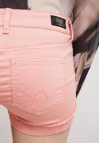 I pantaloni di jeans rosa presentano un orlo sfilacciato, una singola tasca posteriore con un motivo decorativo di cuciture e un'etichetta del marchio sulla cintura.