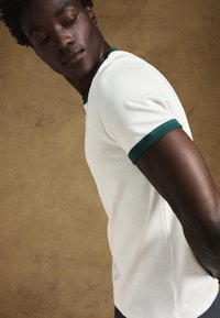 Biała bawełniana koszulka z zielonym prążkowanym kołnierzem i krawędziami rękawów, lekko elastyczna i dopasowanego kroju, na tle o fakturze.